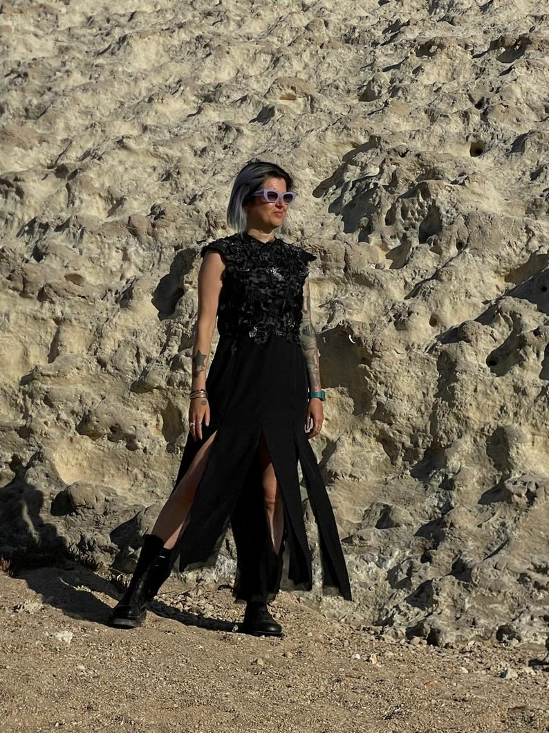 Puede incluir: Un vestido negro sin mangas con un corpi&ntilde;o texturizado y adornado con aplicaciones florales, y una falda larga y fluida con dos aberturas frontales prominentes. La prenda se combina con botas negras con cordones y gafas de sol claras.