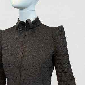 May include: A dark brown long-sleeved jacket featuring a high collar with a front zipper closure. The fabric has a textured, embossed pattern throughout, and the shoulders are structured with a pointed, angular design.