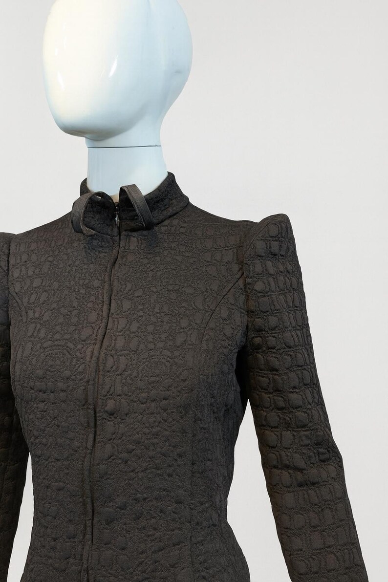 May include: A dark brown long-sleeved jacket featuring a high collar with a front zipper closure. The fabric has a textured, embossed pattern throughout, and the shoulders are structured with a pointed, angular design.