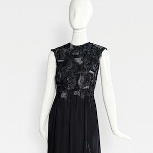 Puede incluir: Un vestido negro sin mangas hasta el suelo con cuello redondo y un corpi&ntilde;o adornado con aplicaciones de tela texturizada en capas. La falda est&aacute; hecha de un material negro liso y s&oacute;lido que cae en una silueta recta y fluida.