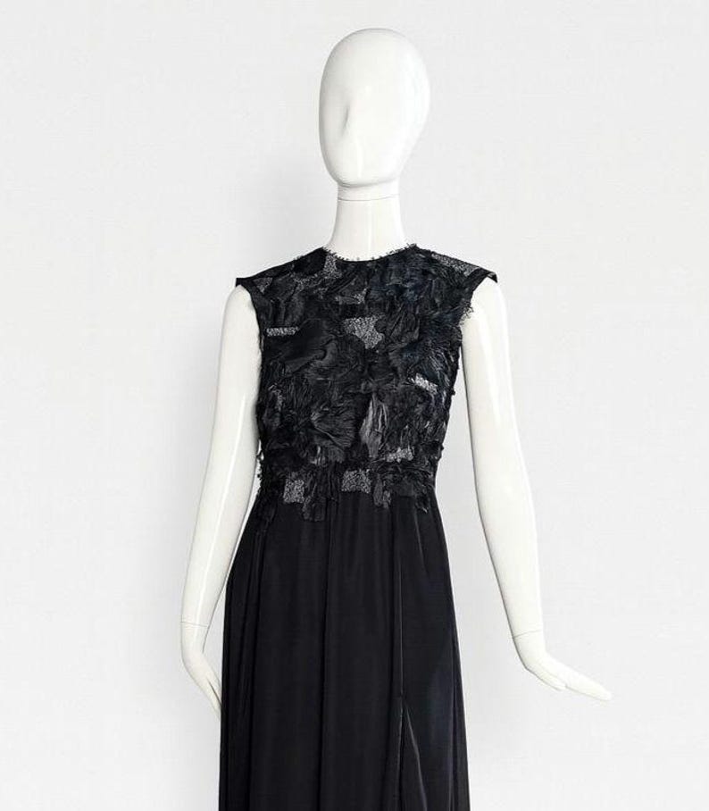 Puede incluir: Un vestido negro sin mangas hasta el suelo con cuello redondo y un corpi&ntilde;o adornado con aplicaciones de tela texturizada en capas. La falda est&aacute; hecha de un material negro liso y s&oacute;lido que cae en una silueta recta y fluida.