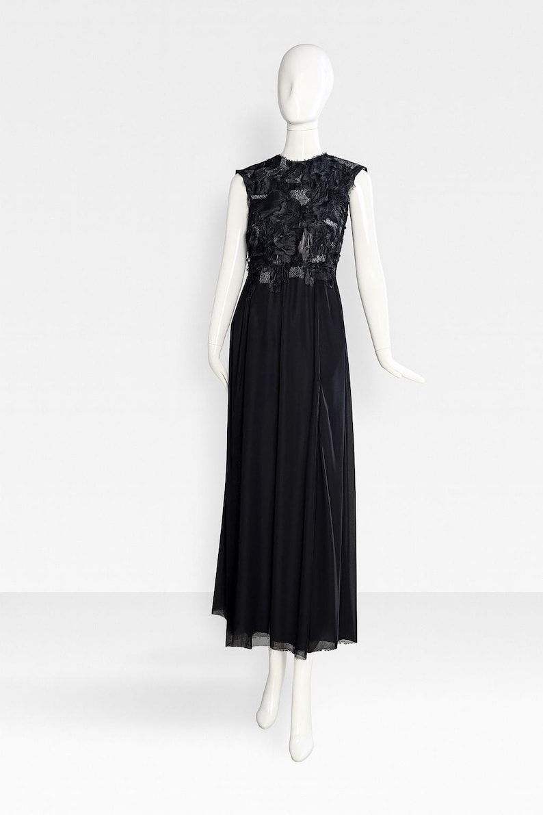 Puede incluir: Un vestido largo negro sin mangas con un corpi&ntilde;o adornado con aplicaciones florales de tela en capas y una superposici&oacute;n transparente. La falda est&aacute; hecha de una tela negra s&oacute;lida que cae hasta el suelo, creando una silueta larga y fluida.