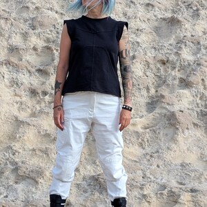 May include: A black sleeveless top with structured shoulders and visible seam detailing paired with white cargo-style pants and black mid-calf boots featuring multiple horizontal straps and buckles on the front.