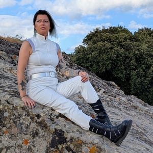 May include: A white sleeveless jumpsuit with a high collar, light blue shoulder accents, and a silver belt, paired with black mid-calf boots featuring multiple horizontal straps and cutouts, worn by a person with visible arm tattoos.