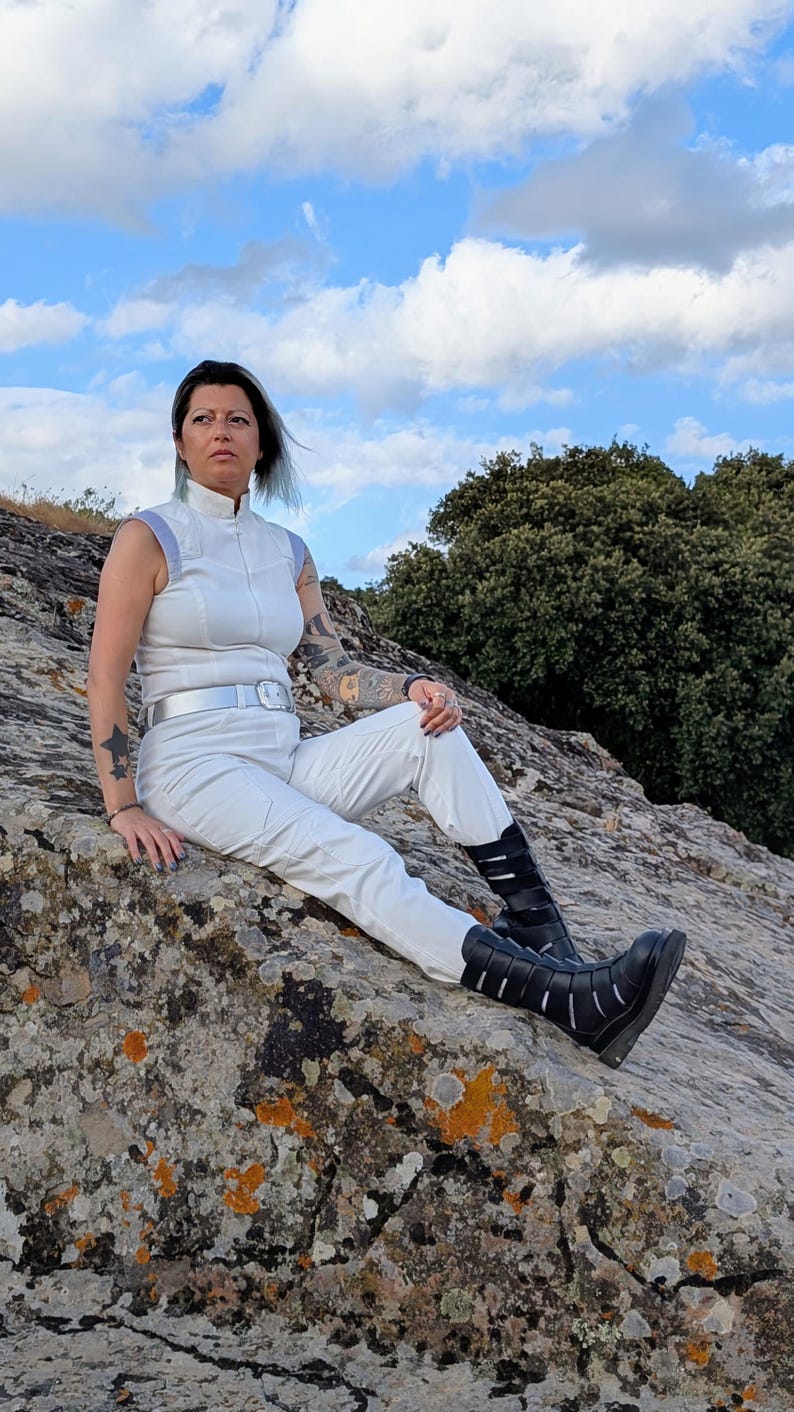 May include: A white sleeveless jumpsuit with a high collar, light blue shoulder accents, and a silver belt, paired with black mid-calf boots featuring multiple horizontal straps and cutouts, worn by a person with visible arm tattoos.