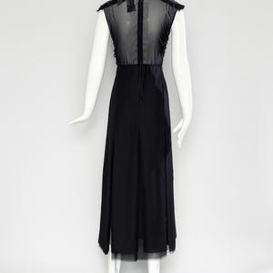 Puede incluir: Un vestido midi negro sin mangas con un panel superior de malla transparente en la espalda, una falda opaca s&oacute;lida y una cremallera vertical en el centro de la espalda. La prenda se muestra sobre un maniqu&iacute; blanco con fondo claro.