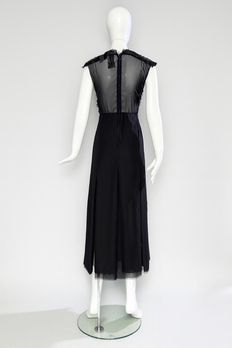 Puede incluir: Un vestido midi negro sin mangas con un panel superior de malla transparente en la espalda, una falda opaca s&oacute;lida y una cremallera vertical en el centro de la espalda. La prenda se muestra sobre un maniqu&iacute; blanco con fondo claro.
