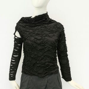 May include: A black long-sleeve top featuring a high neckline and a heavily distressed, shredded texture throughout the fabric. The right sleeve is detached at the shoulder, revealing a cutout, while the left sleeve remains fully attached.