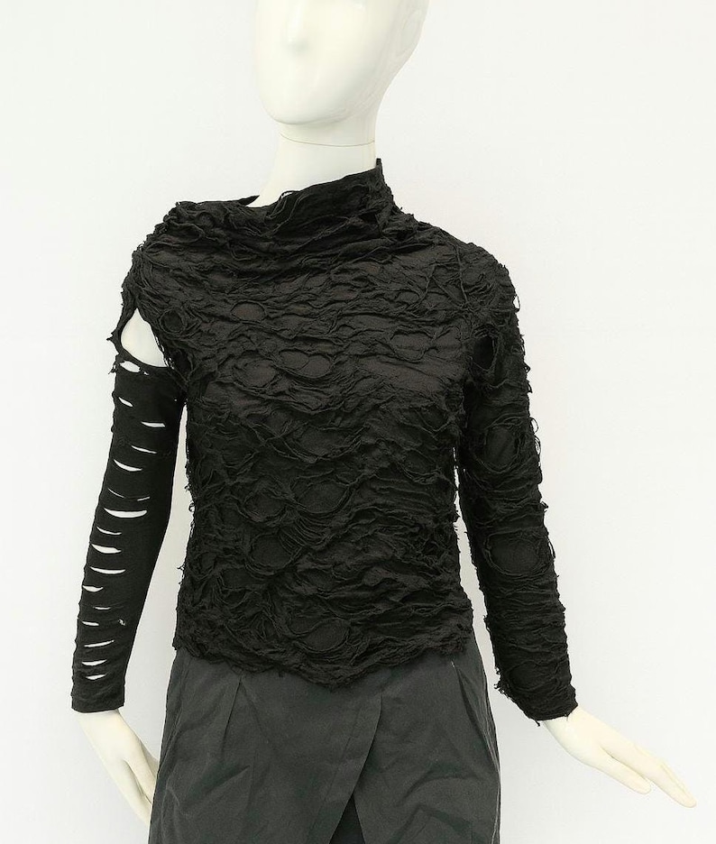 May include: A black long-sleeve top featuring a high neckline and a heavily distressed, shredded texture throughout the fabric. The right sleeve is detached at the shoulder, revealing a cutout, while the left sleeve remains fully attached.