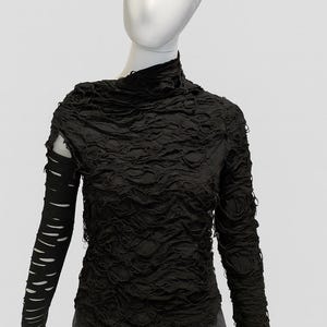May include: A black long-sleeve top featuring a high neckline and a heavily distressed, shredded texture throughout the bodice. The right sleeve is solid black with horizontal cutouts, while the left sleeve matches the shredded body.