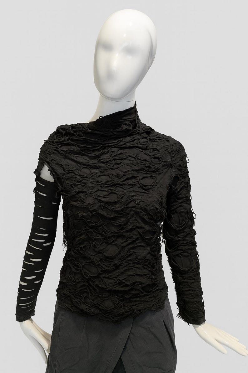 May include: A black long-sleeve top featuring a high neckline and a heavily distressed, shredded texture throughout the bodice. The right sleeve is solid black with horizontal cutouts, while the left sleeve matches the shredded body.