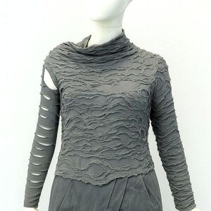 May include: A charcoal gray long-sleeve top with a high neckline and a textured, crinkled fabric surface. The right sleeve features a series of horizontal cutouts along the arm, while the left sleeve remains solid. The garment has a fitted silhouette.