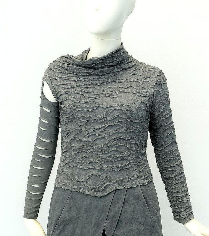 May include: A charcoal gray long-sleeve top with a high neckline and a textured, crinkled fabric surface. The right sleeve features a series of horizontal cutouts along the arm, while the left sleeve remains solid. The garment has a fitted silhouette.