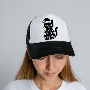 May include: A black and white trucker hat featuring a grumpy cat wearing a Santa hat and wrapped in Christmas lights. The hat has a black brim and mesh back, with a white front panel.