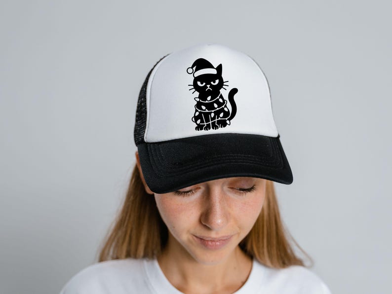 May include: A black and white trucker hat featuring a grumpy cat wearing a Santa hat and wrapped in Christmas lights. The hat has a black brim and mesh back, with a white front panel.