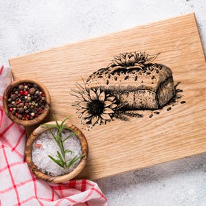 Puede incluir: Una tabla de cortar de madera con una ilustración en blanco y negro de una hogaza de pan, girasoles y trigo. Dos cuencos de madera pequeños contienen granos de pimienta y sal con romero. Un paño a cuadros rojo y blanco es parcialmente visible.