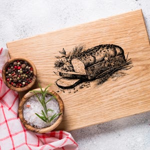 Könnte beinhalten: Ein hölzernes Schneidebrett mit einer detaillierten Illustration eines Brotlaibs, geschnitten, mit Weizen- und Blumenelementen. Zwei kleine Holzschalen enthalten Pfefferkörner und Salz mit Rosmarin sowie ein rot-weiß kariertes Tuch.