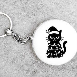 May include: A round white keychain with a silver chain and ring. The keychain features a black illustration of a grumpy cat wearing a Santa hat and wrapped in Christmas lights. The cat has an angry expression.