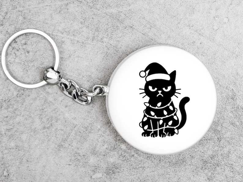 May include: A round white keychain with a silver chain and ring. The keychain features a black illustration of a grumpy cat wearing a Santa hat and wrapped in Christmas lights. The cat has an angry expression.