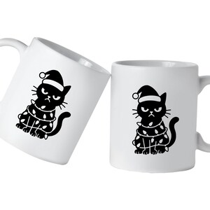 May include: Two white ceramic mugs with a black graphic of a grumpy cat wearing a Santa hat and wrapped in Christmas lights. The cat has an angry expression. One mug is tilted.