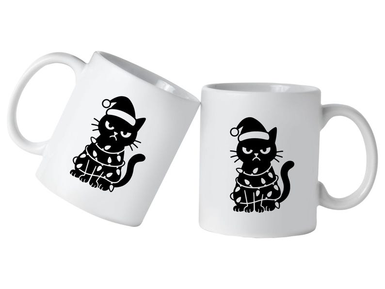 May include: Two white ceramic mugs with a black graphic of a grumpy cat wearing a Santa hat and wrapped in Christmas lights. The cat has an angry expression. One mug is tilted.