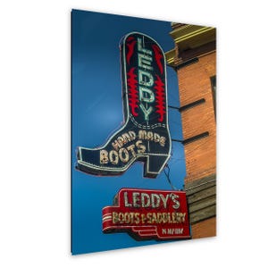 May include: A neon sign in the shape of a cowboy boot with the text "LEDDY'S HAND MADE BOOTS" against a blue sky. Below, a red sign reads "LEDDY'S BOOTS & SADDLERY N. MAIN". The signs are mounted on a brick building.