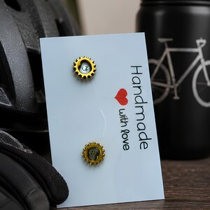 May include: Two gold-colored gear-shaped stud earrings attached to a white display card. The card features the text "Handmade with love" in black lettering, accompanied by a small red heart icon positioned between the two lines of text.