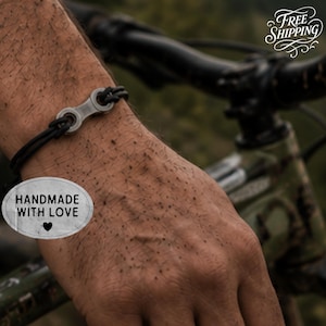 May include: A bracelet featuring a silver-toned metal bicycle chain link connector attached to a thin black cord. The bracelet is displayed on a wrist, with a small oval sticker visible that reads HANDMADE WITH LOVE and a heart symbol.