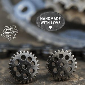 May include: A pair of metallic, dark grey gear-shaped stud earrings with a circular design featuring multiple small cut-out holes. The earrings have a textured, industrial appearance and are positioned in front of a blurred bicycle chain.
