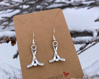 May include: Silver hockey stick earrings with a hook closure, displayed on a brown card with the text "Handmade with love" and a red heart. The earrings are set against a snowy, wintery background.