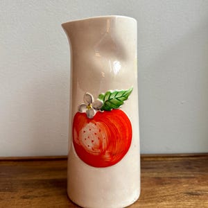 Puede incluir: Una jarra de cerámica de color crema con cuerpo cilíndrico y pico moldeado. La parte frontal presenta un diseño en relieve pintado a mano de una manzana roja brillante con una pequeña flor blanca y una hoja verde en la parte superior.