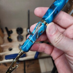 May include: A handcrafted pen clip with a vibrant blue and brown marbled resin barrel. The pen clip features gold-coloured metal accents and a functional clip. This unique writing accessory combines artistry and utility.