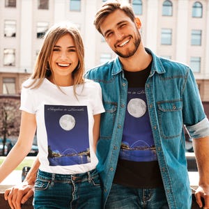 May include: White t-shirt with the text "Sleepless in Newcastle" above a graphic of a full moon over a city skyline. The t-shirt is worn by a person. The other person is wearing a black t-shirt with the same graphic.