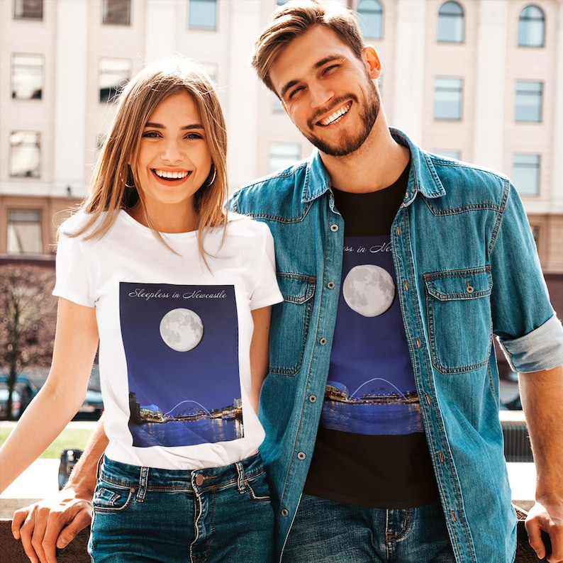 May include: White t-shirt with the text "Sleepless in Newcastle" above a graphic of a full moon over a city skyline. The t-shirt is worn by a person. The other person is wearing a black t-shirt with the same graphic.