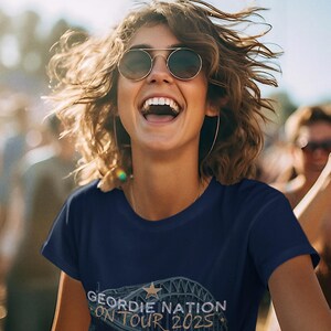 May include: A navy blue t-shirt featuring the text "GEORDIE NATION ON TOUR 2025". The design includes a star and a bridge illustration. The image shows a person wearing sunglasses and smiling.