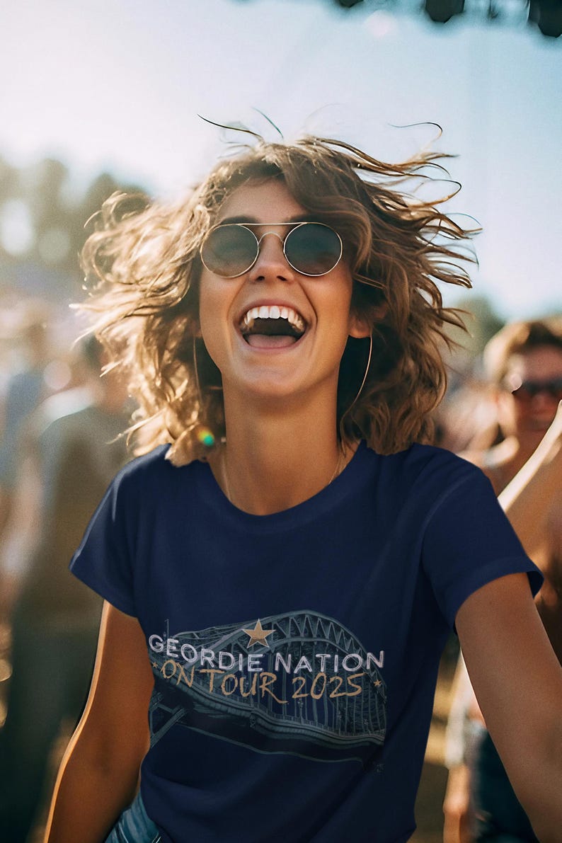 May include: A navy blue t-shirt featuring the text "GEORDIE NATION ON TOUR 2025". The design includes a star and a bridge illustration. The image shows a person wearing sunglasses and smiling.