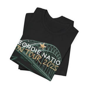 May include: Black t-shirt with a graphic of a bridge, a star, and the text "Geordie Nation On Tour 2025" in white and gold. The shirt is folded. The design suggests a tour or event.