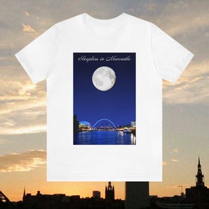 May include: White t-shirt featuring a nighttime scene with a full moon over a city skyline and a bridge. The text "Sleepless in Newcastle" is printed above the image. The shirt has short sleeves and a crew neck.