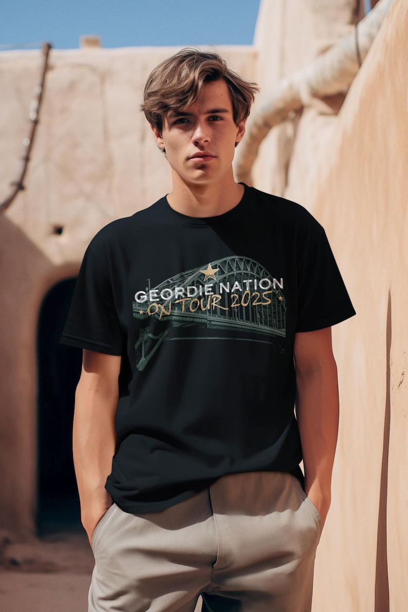 May include: Black t-shirt with the text "GEORDIE NATION ON TOUR 2025" in white and gold. The design features a bridge graphic with a star. The shirt is worn by a person.