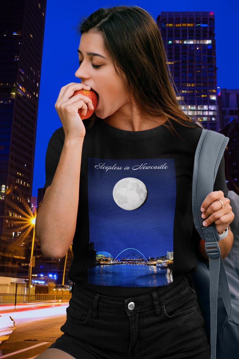 May include: Black t-shirt with a graphic of a full moon over a city skyline and the text "Sleepless in Newcastle." The person is eating an apple. The background shows a city at night.
