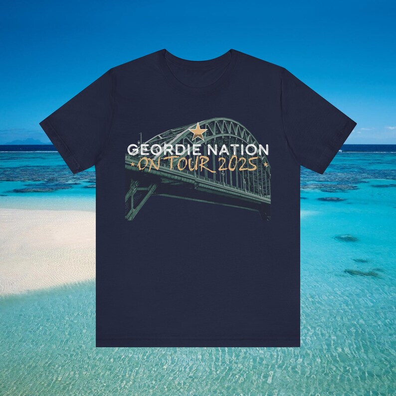 May include: A navy blue t-shirt with the words "GEORDIE NATION ON TOUR 2025" printed on it, along with an image of a bridge. The text is in white and gold. The background shows a beach and the sea.