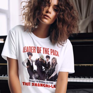 Puede incluir: Camiseta blanca con el texto rojo "LEADER OF THE PACK". Debajo, una imagen en blanco y negro de una banda en una motocicleta. El texto "He came from the wrong side of town THE SHANGRI-LA" está impreso debajo.