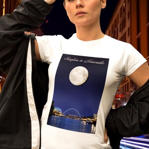 May include: White t-shirt with a graphic of a full moon over a city skyline and the text "Sleepless in Newcastle." The shirt is worn with a black jacket and sunglasses.
