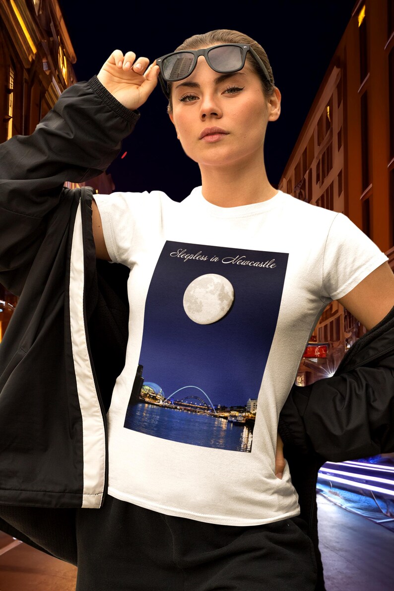 May include: White t-shirt with a graphic of a full moon over a city skyline and the text "Sleepless in Newcastle." The shirt is worn with a black jacket and sunglasses.