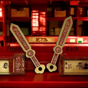 May include: Two decorative gold-colored swords with red accents and Chinese characters. The swords have a unique shape with a circular design and a hole at the base of the handle. The background is a red-toned interior.