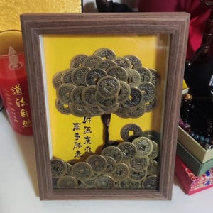 Könnte beinhalten: Ein gerahmtes Kunstwerk mit einem Baum, der aus zahlreichen bronzefarbenen Münzen gefertigt wurde. Der Baum ist vor einem gelben Hintergrund mit chinesischen Schriftzeichen platziert. Der Rahmen ist aus Holz.