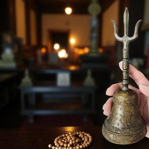 May include: A bronze-colored bell with a trident-shaped handle is held in front of a blurred background. The bell has intricate carvings and a round base. A wooden bead necklace rests on a dark wooden surface.