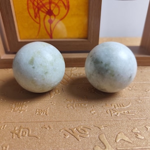 May include: Two pale green marble spheres with subtle veining. The spheres are smooth and round, resting on a textured, golden-brown surface. A wooden picture frame and a small figurine are in the background.