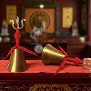 Puede incluir: Dos campanas de latón con asas de tridente y detalles de tela roja descansan sobre una tela roja. Una campana está de pie, mientras que la otra está acostada. Volutas de humo se elevan.