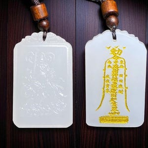 Könnte beinhalten: Zwei weiße Jade-Anhänger mit aufwendigen Schnitzereien. Einer zeigt ein detailliertes Relief einer Figur, der andere goldfarbene chinesische Schriftzeichen. Jeder Anhänger ist rechteckig mit einem gewellten Oberteil, auf einer braunen Kordel.